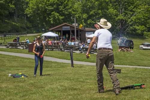 A man in a cowboy hat throws a bean bag while a woman prepares to catch it, with a gathering in the background.