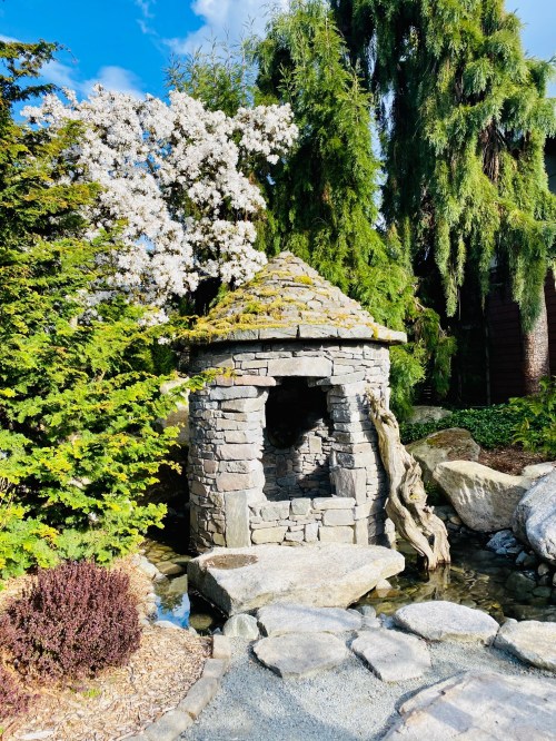 A stone structure with a mossy roof surrounded by greenery and flowering trees, near a small pond.