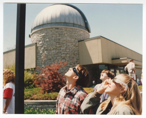 A group of people wearing sunglasses look up at the sky near a dome-shaped observatory building.