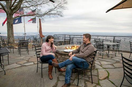 A couple sits by a fire pit on a patio, enjoying a scenic view with flags in the background.