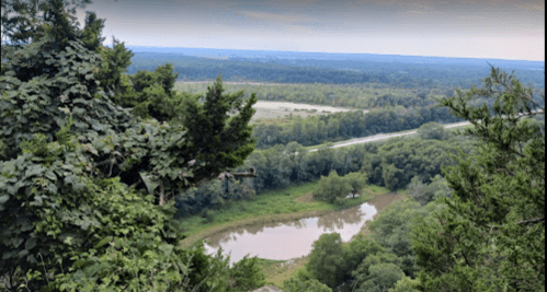 A scenic view from a high vantage point, overlooking a river and lush green landscape under a cloudy sky.