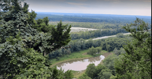 A scenic view from a high vantage point, overlooking a lush landscape with a winding road and a calm pond.