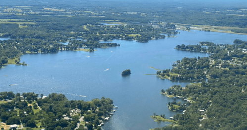 Aerial view of a serene lake surrounded by greenery, with small islands and a distant industrial area.