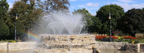 A stone fountain sprays water in a park, with trees and colorful flowers in the background under a blue sky.