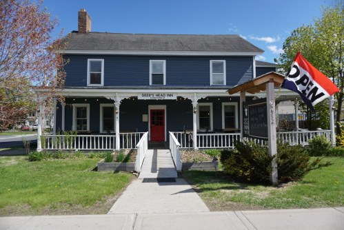 A blue inn with a red door and an "OPEN" flag, surrounded by greenery and a clear blue sky.