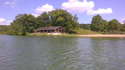 A serene lakeside view featuring a cabin surrounded by trees and a sandy beach under a blue sky with fluffy clouds.