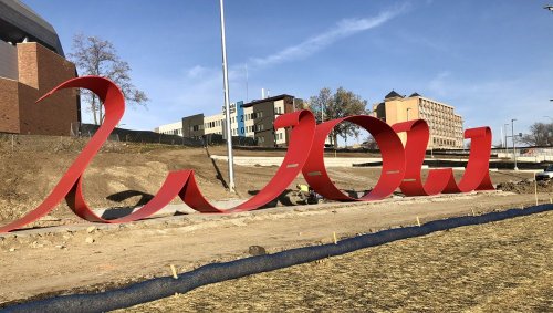 A large red sculpture spelling "wow" in an outdoor setting, with buildings in the background and construction elements nearby.