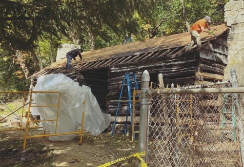 Two workers repair a rustic log cabin roof, with scaffolding and a fence in the foreground, surrounded by trees.