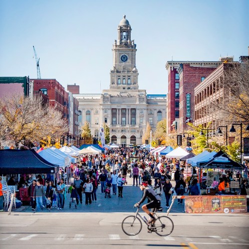 A bustling street market with tents and crowds, set against a historic building with a clock tower.