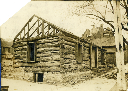 A partially constructed log cabin with a wooden frame, set in a rural area with trees and nearby buildings.