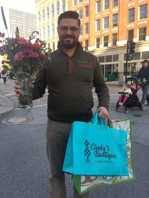 A man holds a bouquet of flowers and shopping bags while standing on a city street.