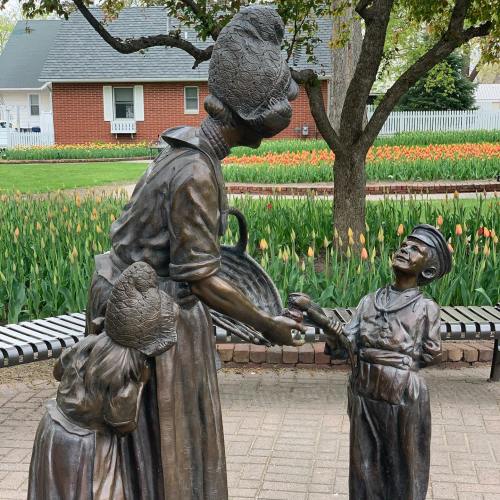 A bronze statue of a woman handing a basket to a boy, with a girl leaning against her, set in a garden with tulips.