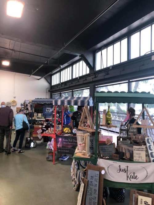A vibrant market scene with vendors displaying handmade goods and shoppers browsing in a spacious indoor setting.