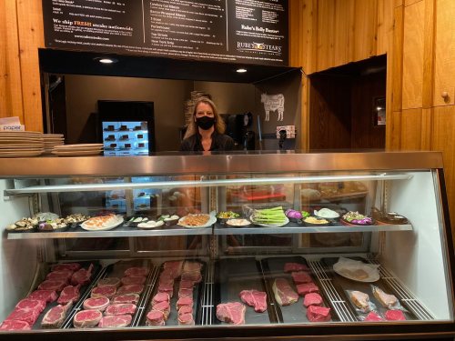 A woman wearing a mask stands behind a display case filled with various meats and seafood in a butcher shop.