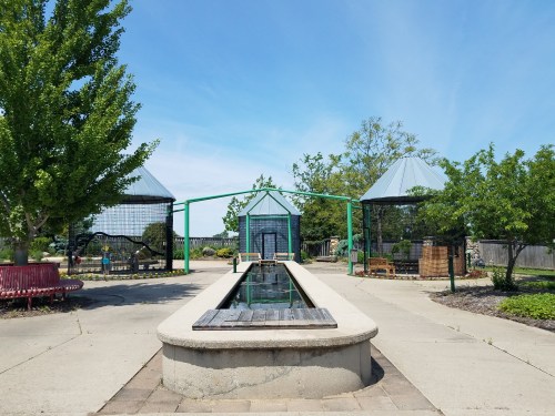 A serene garden with a central water feature, surrounded by green trees and two gazebo-like structures under a clear blue sky.