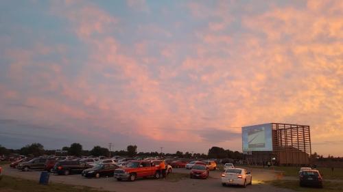 A drive-in movie theater at sunset, with cars parked in front of a large screen and colorful clouds in the sky.