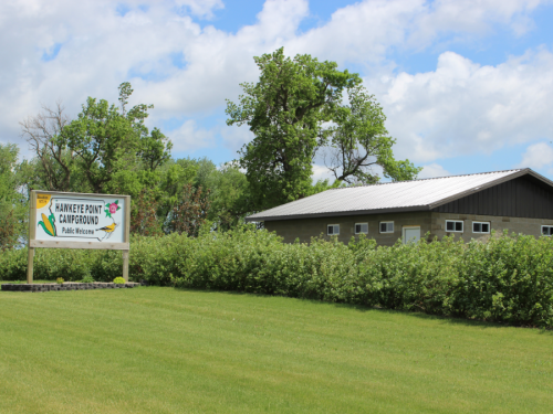Sign for Hawkeye Point Campground with a public restroom building in the background, surrounded by greenery and blue skies.