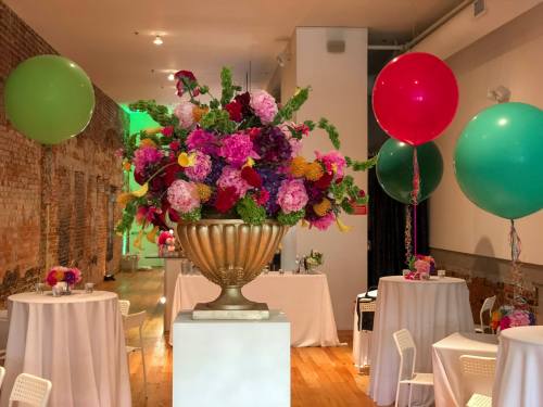 A vibrant floral arrangement in a gold vase, surrounded by tables and large balloons in green and pink.