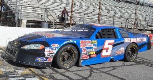 A blue NASCAR race car with the number 5, parked on a racetrack with grandstands in the background.