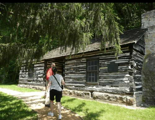 Two people walk towards a rustic log cabin surrounded by greenery and trees on a sunny day.