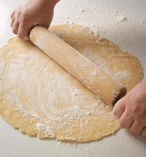 Hands using a rolling pin to flatten dough on a floured surface.