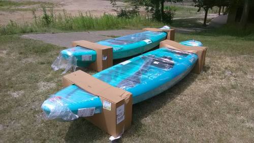 Two wrapped kayaks in cardboard boxes, resting on grass near a pathway.