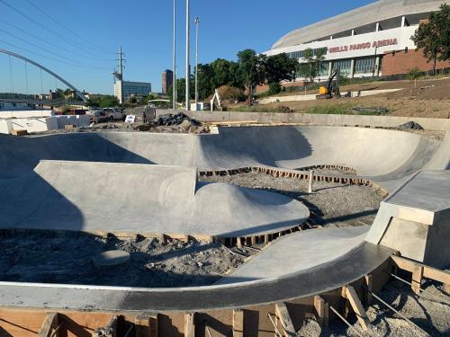 A partially completed concrete skatepark with various ramps and curves, set against a backdrop of a sports arena.