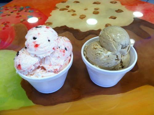 Two cups of ice cream: one with pink and white swirls and chocolate bits, the other a light brown flavor, on a colorful table.