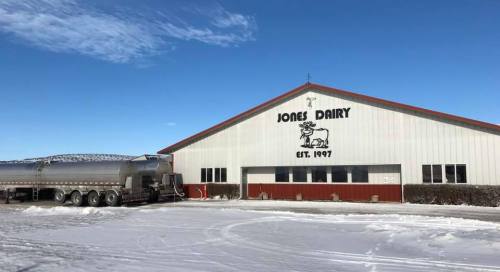 A dairy farm building with a truck parked outside, snow covering the ground, and a clear blue sky above.