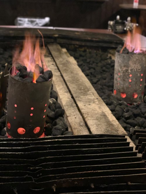 Two charcoal chimneys with glowing embers, surrounded by black stones, set in a rustic indoor setting.