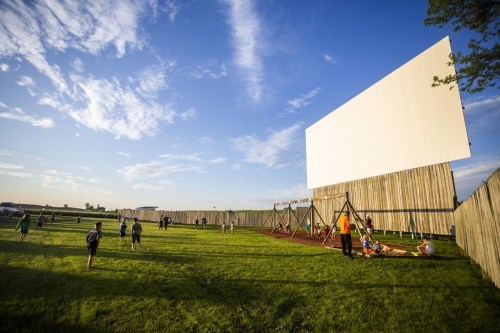 A grassy area with a large blank screen, playground equipment, and people enjoying the outdoors under a blue sky.