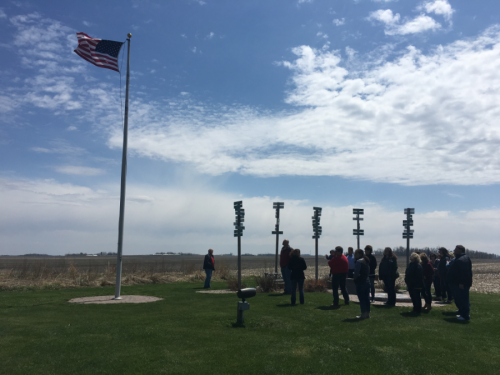 A group of people stands in a field, observing a flagpole with an American flag and several markers in the background.