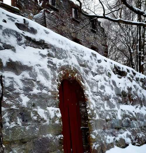 A snow-covered stone wall with a red door, leading to a historic building partially obscured by trees.