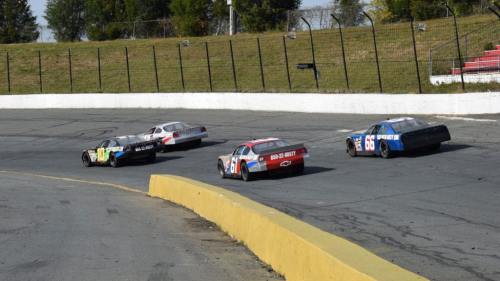 Four race cars compete on a circular track, with a grassy area and bleachers in the background.