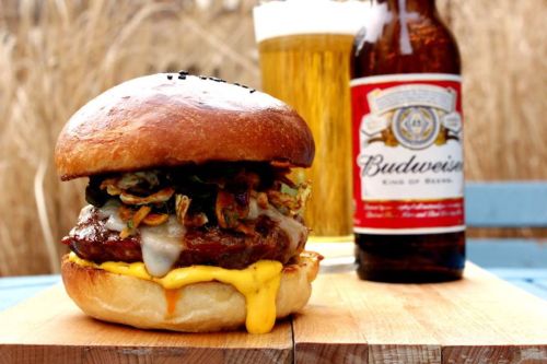A juicy burger with melted cheese and toppings, alongside a bottle of Budweiser and a glass of beer.