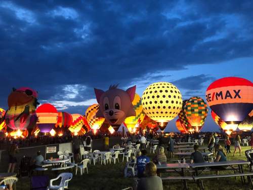 Colorful hot air balloons illuminated at dusk, with spectators seated at tables enjoying the vibrant display.