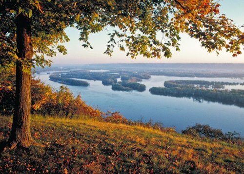 A scenic view of a river winding through autumn-colored trees and hills under a clear sky.