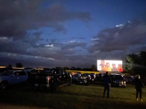 A drive-in movie scene at dusk, with cars parked and a large screen displaying a film against a cloudy sky.