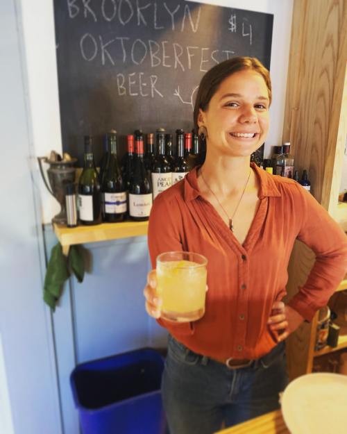 A smiling woman in a red shirt holds a drink at a bar with wine bottles and a chalkboard menu in the background.