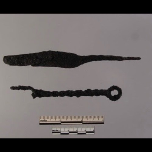 Two ancient iron artifacts, one elongated and pointed, the other twisted with a circular end, displayed with a ruler for scale.