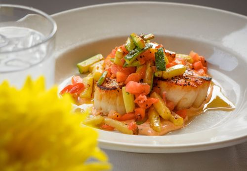 A plate of seared scallops topped with diced vegetables, served with a creamy sauce and a glass of water in the background.