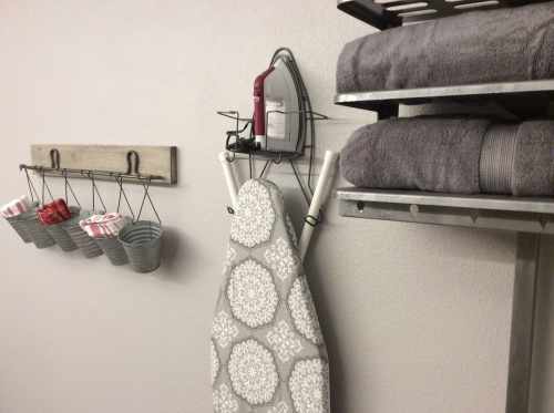 An ironing board with a gray cover, an iron hanging above, and neatly stacked towels on a shelf beside hanging baskets.