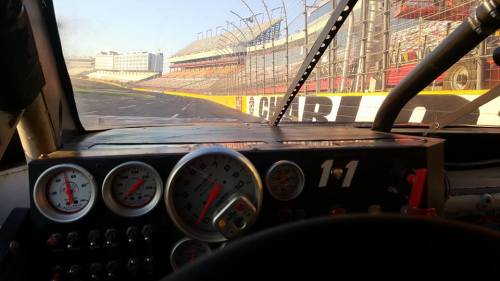 View from inside a race car, showing the dashboard with gauges and a racetrack in the background.