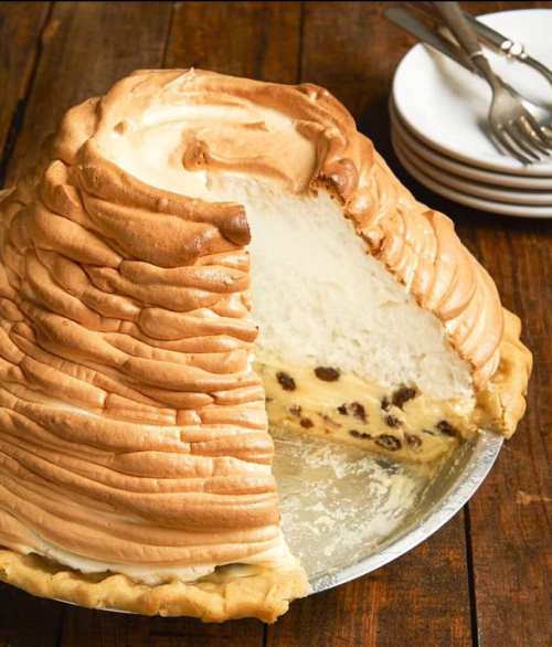 A large, fluffy meringue pie with a slice cut out, revealing a creamy filling and raisins, on a wooden table.