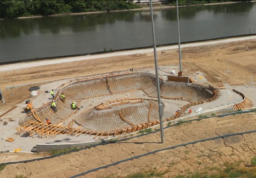 Construction site with workers assembling a concrete structure near a river, surrounded by wooden forms and rebar.
