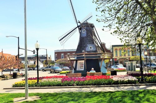 A colorful flower bed surrounds a wooden windmill in a sunny park, with benches for visitors to relax.