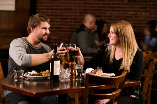 A couple clinks wine glasses at a cozy restaurant, enjoying a meal together. Warm atmosphere with brick walls in the background.