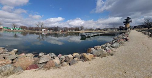 A serene lakeside view with rocky shores, docks, and a cloudy sky reflecting on the water's surface.