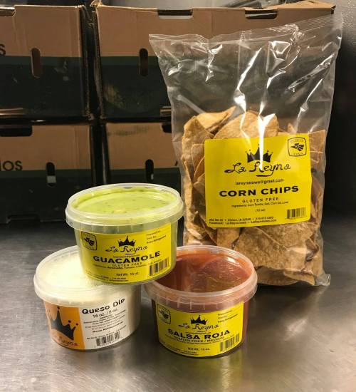 Three containers of dips (guacamole, queso dip, salsa roja) and a bag of corn chips on a countertop, with boxes in the background.