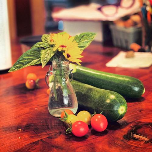 A small glass vase with yellow flowers sits on a wooden table, surrounded by zucchini and colorful cherry tomatoes.